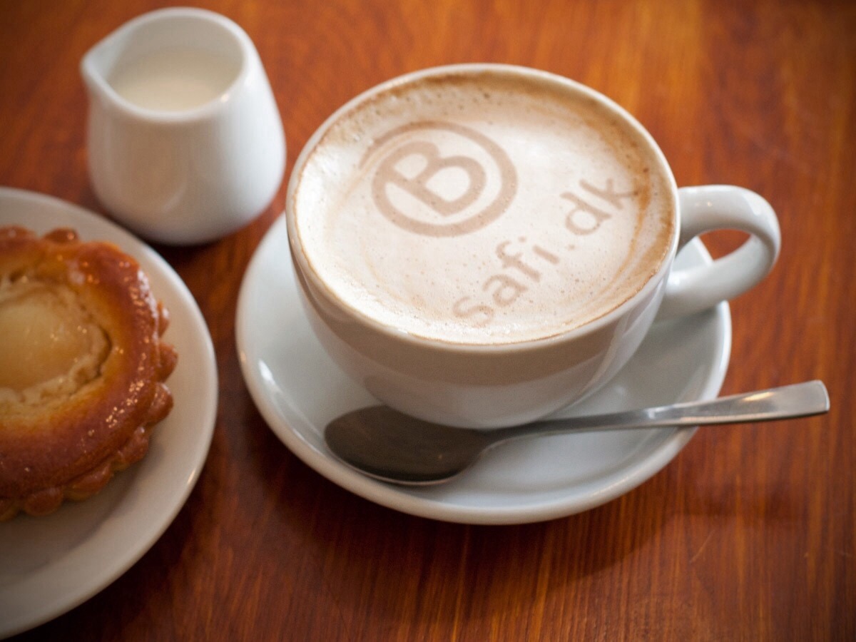 A cup of coffee with SAFI.DK written in the foam, and a pastry on a table.